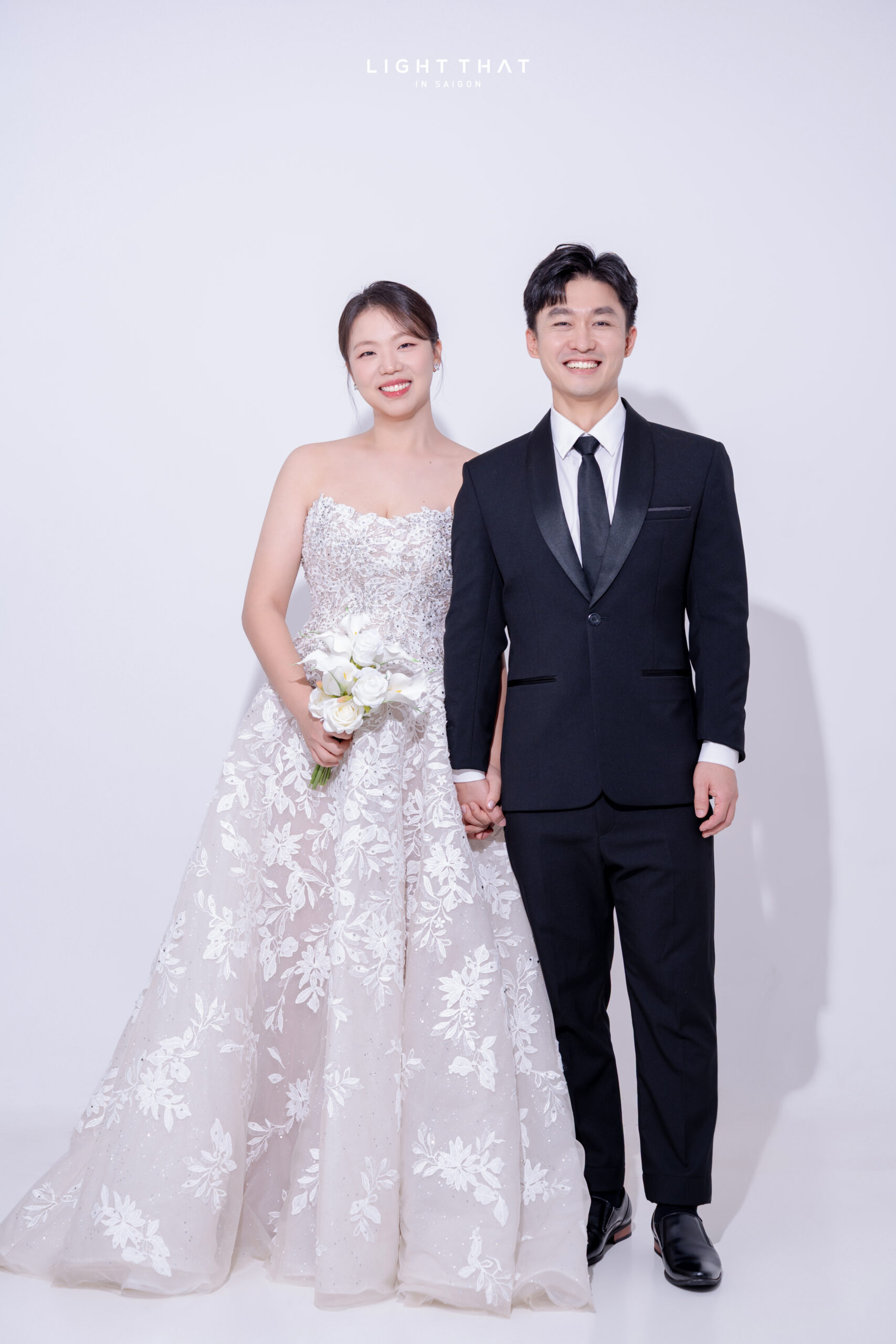 Cô dâu chú rể tạo dáng đơn giản trước phông trơn trắng tại studio chụp ảnh cưới, nụ cười tự nhiên và ánh sáng dịu